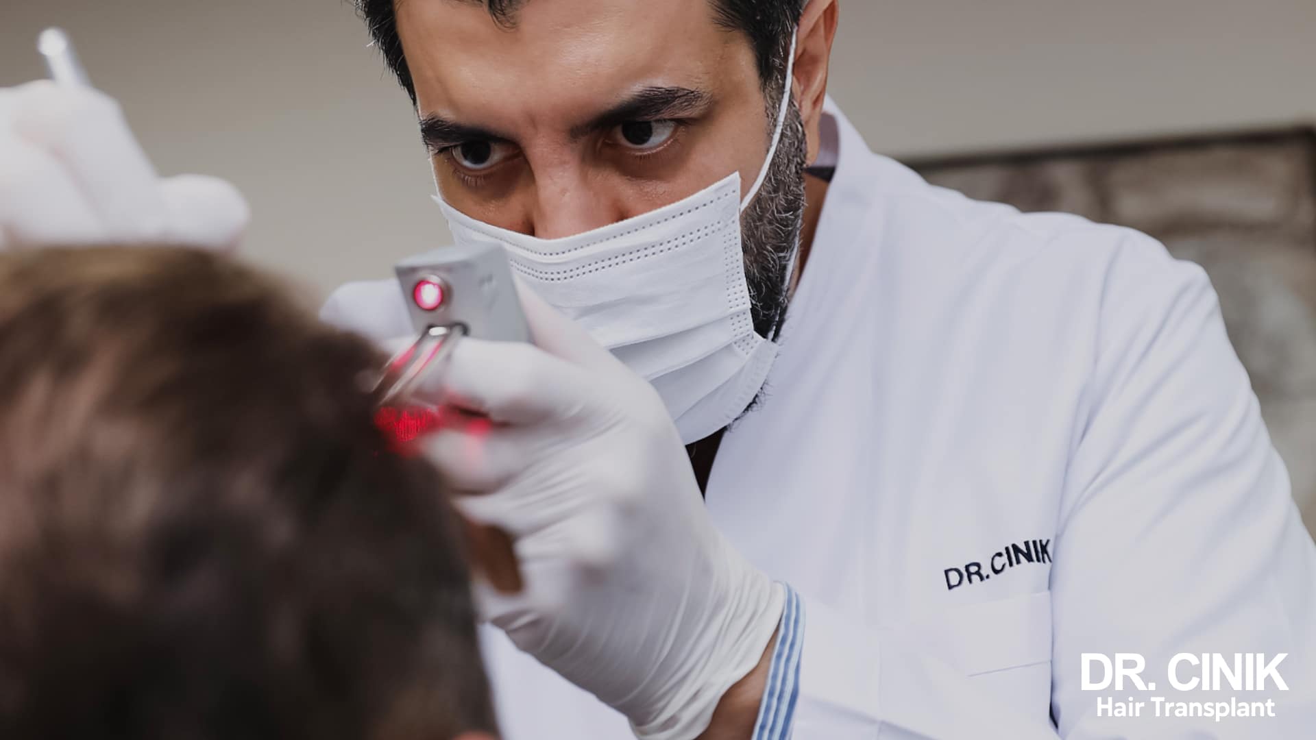 Il dottor Emrah Cinik, con mascherina e camice bianco, osserva da vicino il cuoio capelluto di un paziente con uno strumento di ingrandimento a luce rossa
